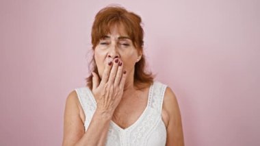 Tükenmiş orta yaşlı kadın sürekli esniyor; tembellik izole edilmiş pembe arka planda yakalanmış. Sıkıntı, yorgun ifadelerin her yönüyle görülebilir ve elleri ağızlarını kapatır..