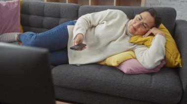 Orta yaşlı İspanyol kadın, renkli yastıklarla bir kanepede uzanıyor, elinde televizyon kumandası var, evinde rahat ve memnun.