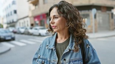 Kıvırcık saçlı, güneş gözlüğü takan ve kot ceket giyen orta yaşlı bir kadın şehir yolunda geziniyor.