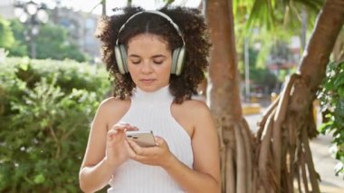 Kıvırcık saçlı genç bir İspanyol kadın şehir parkında kulaklıklarıyla müzik dinliyor..