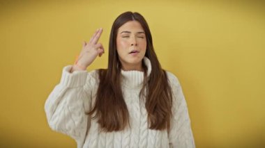 Paniğe kapılmış genç İspanyol kadın intihar süsü veriyor, parmakları kafaya dayalı silah oluşturuyor, sarı izole arka planda duruyor..