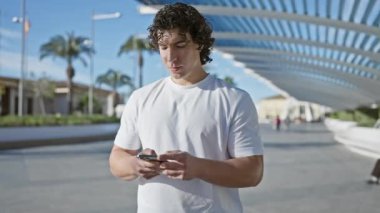 Gülümseyen genç İspanyol adam arka planında modern mimari ve palmiye ağaçları olan akıllı telefon kullanıyor..