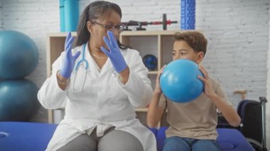 Bir kadın terapist klinikte mavi taşak kullanan bir çocukla rehabilitasyon egzersizi yapıyor.