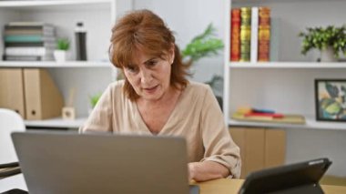 Neşeli orta yaşlı kadın kendini güvenle sunuyor, kameraya bakarken iki eliyle yüzünü gösteriyor, neşeyle gülümsüyor, bilgisayarla ofiste oturuyor..