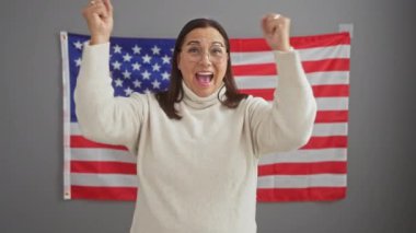 Heyecanlı orta yaşlı İspanyol kadın, gözlük takıyor, ofisteki zaferle dolup taşıyor, gururla duruyor ve bağırıyor, zafer içinde kolunu kaldırıyor, ABD bayrağı yakınındaki başarısını kutluyor.