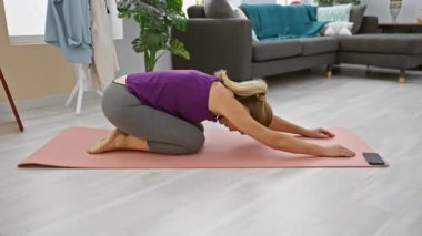 Orta yaşlı bir kadın modern bir oturma odasında pembe bir paspasın üzerinde yoga pozu veriyor. Sağlık ve refahı gösteriyor..
