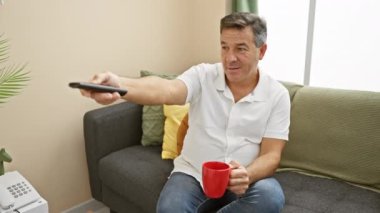 Rahat giyinmiş olgun bir adam. Elinde bir kupa ve uzaktan kumandayla rahat bir oturma odasında oturuyor..