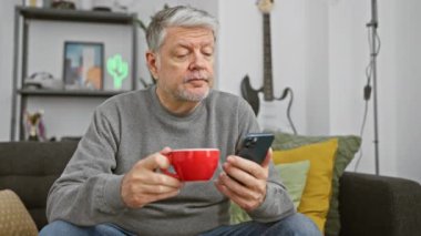 Kahve içen ve oturma odasında akıllı telefon kullanan olgun, gri saçlı bir adam..