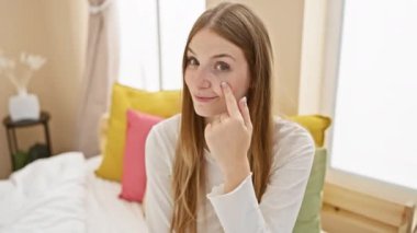 Şüpheli bir ifade, pijamalı genç sarışın kadın, yatak odasından seni dikkatle izliyor, 'seni görüyorum' hareketinde onun gözünü işaret ediyor.