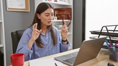 Ofiste başarıyı kutlayan mutlu genç İspanyol kadın. Masasında dizüstü bilgisayarı, kupası ve evrakları var..