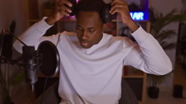 Beyaz kazaklı, odaklanmış bir Afrikalı Amerikalı gece vakti neon ışıklı bir ev stüdyosunda kulaklıklarını ayarlıyor..