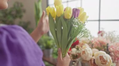 Bir kadın, parlak botanik arka planı olan bir çiçekçide sarı ve mor laleleri inceliyor..