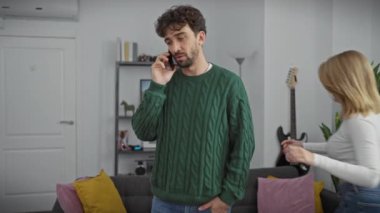 Oyuncu bir kadın, modern bir oturma odasında telefonla konuşurken odaklanmış erkeğini sırtında taşır..