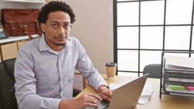 Ciddi bir iş! Yakışıklı, Afro-Amerikan bir adam, ofiste havalı bir stil işçisi, masada otururken kameraya doğal ve basit bir ifadeyle bakıyor..