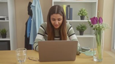 Gülümseyen genç bir İspanyol kadın evde dizüstü bilgisayarda çalışıyor, modern bir iç mekan ortamında rahat ve çekici bir yetişkini canlandırıyor..