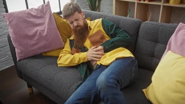 Bearded Man Colorful Shirt Experiences Discomfort While Sitting Grey Sofa — Stock Video