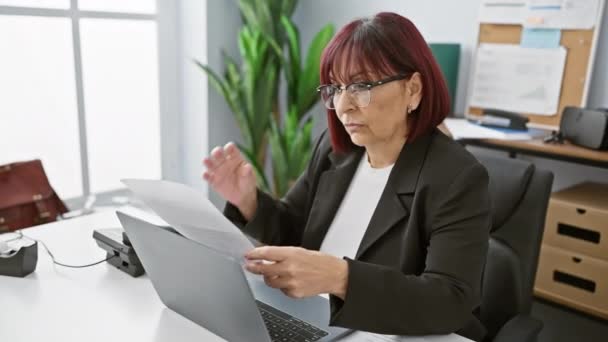 一位忧心忡忡的中年妇女在室内的办公室里拿起眼镜 手里拿着文件 坐在笔记本电脑旁 — 图库视频影像