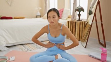 Sağlıksız genç Çinli kadın mide ağrısı çekiyor, mide bulantısı hastalığıyla mücadele ediyor, yoga minderinin üstünde oturuyor, yatak odasında rahatsızlığını dile getiriyor..