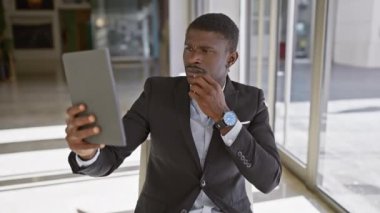 Takım elbiseli Afrikalı adam modern ofis odasında tabletle selfie çekiyor.