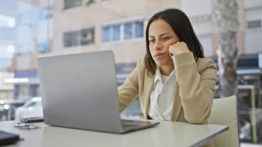 Genç İspanyol bir kadın laptopuyla çalışırken yorgun ve stresli hissediyor. Geçmişi bulanık ve modern bir ofiste..