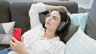 Rahat genç bir kadın, modern oturma odasındaki bir kanepede yatarken kulaklıkla müzik dinlemekten hoşlanır..