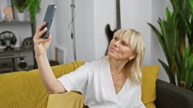 Neşeli, olgun, beyaz bir kadın modern dekorlu rahat bir oturma odasında selfie çekiyor..