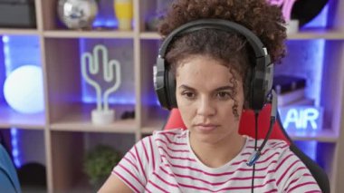 Ciddi yüzlü İspanyol kadın, doğal, basit görünüşlü ve kıvırcık saçlı, yoğun bir şekilde loş ışıklı oyun odasındaki kameraya bakıyor..