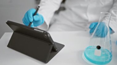 Laboratuvar önlüklü bir bilim adamı laboratuarda tablet kullanırken mavi bir sıvıyı inceliyor.