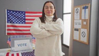 Gülümseyen İspanyol bir kadın, arka planda Amerikan bayrakları ve oy sandığı olan bir Amerikan seçim merkezinde çapraz kollarıyla duruyor..