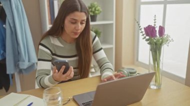 Evde dizüstü bilgisayarı, defteri, çiçekleri ve kulaklığı olan akıllı telefon kullanan genç bir İspanyol kadın var..