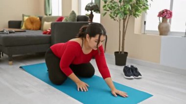 Genç bir İspanyol kadın, modern oturma odasında mavi bir yoga minderine uzanır. Bu da sağlıklı ve aktif bir yaşam tarzını gösterir..