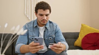 Puzzled man holding credit card and smartphone while sitting on a grey sofa indoors, expressing concern with financial management.