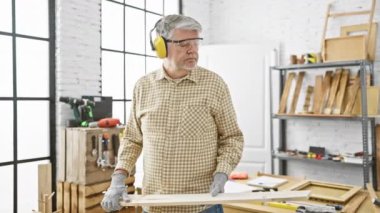 Gözlük takan ve koruyucu kulaklık takan olgun bir adam, iyi aydınlatılmış bir marangozluk atölyesinde kereste tutuyor..