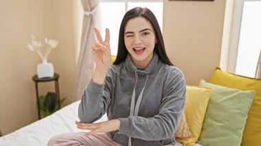 Pijamalı, genç, güzel İspanyol bir kadın, yatak odasında güven içinde oturuyor, kazanan bir gülümseme ve zafer işareti gösteriyor, kendini davetkar bir şekilde canlı hissettiriyor.!
