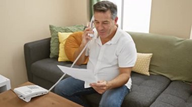 Beyaz gömlekli olgun bir adam, elinde belge tutarken ev telefonuyla konuşuyor..