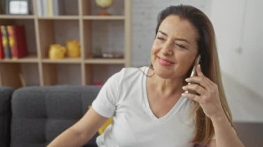 Oturma odasında telefonda konuşan heyecanlı bir kadın, sürpriz ve mutluluğu ifade ediyor, günlük hayatın samimi bir anını yakalıyor..