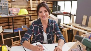 Etrafında aletler ve güvenlik ekipmanlarıyla marangozluk atölyesinde gülen bir kadın..