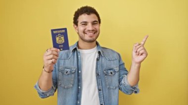 Emirlik pasaportu taşıyan neşeli genç bir Arap adam, sarı izole edilmiş arka planda neşeli bir gülümsemeyle kenarı işaret ediyor.