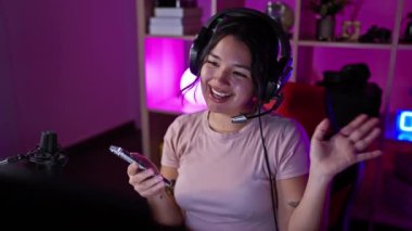 Geceleri neon ışıklı oyun odasında akıllı telefon kullanan, kulaklıklı, gülümseyen bir kadın.