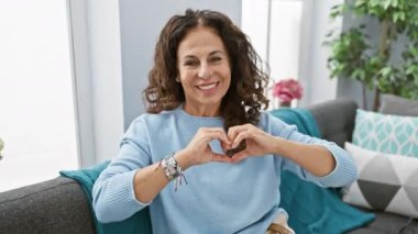 Orta yaşlı İspanyol bir kadının yürek ısıtan portresi, oturma odasındaki kanepede oturuyor, bir el kalbi sembolü yapıyor. Romantik bir konsept. Sevgililer Günü için evde neşeli bir sürpriz sergilemek..