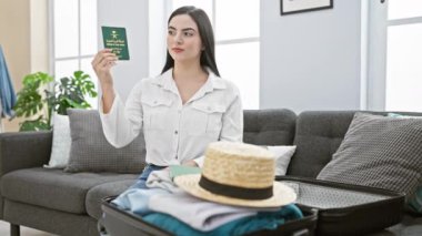 Evdeki neşeli İspanyol kadın, Suudi pasaportu gösteriyor, olumlu başparmak hareketi yapıyor, ağzı açık gülümsüyor, oturuyor ve yan tarafına işaret ediyor.