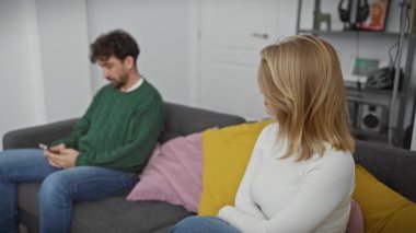 Bir kadın akıllı telefonuna odaklanmış, rahat bir oturma odasındaki kanepede oturan bir adama derin derin bakar..