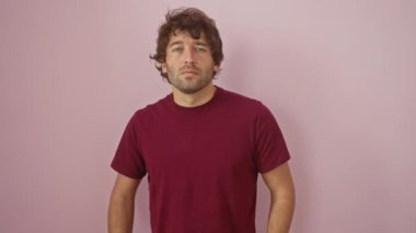 Ciddi görünümlü İspanyol bir adam, basit bir tişört giyiyor, kendine güvenen, doğal ve ciddi duruyor, pembe izole bir arka planda, soğukkanlılıkla kameraya bakıyor