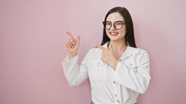 Neşeli genç İspanyol kadın iki eliyle yan yana işaret ediyor, pembe izole edilmiş arka planda güvenli bir şekilde ürünü sunuyor, gülümsüyor, kameraya bakıyor.