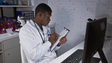 Laboratuvar önlüklü, odaklanmış Afrikalı bir adam modern bir laboratuvar ortamında bilgisayardaki verileri incelerken not alıyor.