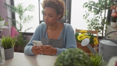 Canlı bitkiler ve bahçe aletleriyle çevrili bir çiçekçide akıllı telefon kullanan Afrikalı Amerikalı kadın..