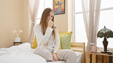 Stresli genç sarışın kadın yatak odasında oturuyor, gergin bir şekilde tırnaklarını yiyor, elleri endişeyle titriyor - pijamalı bir korku portresi gibi görünüyor.