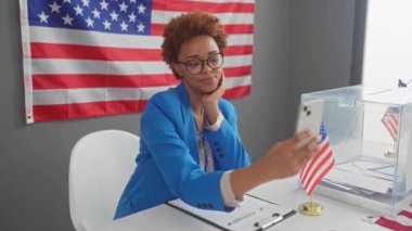 Blazer ceketli Afrikalı Amerikalı kadın telefonuyla selfie çekiyor. Seçmen seçim merkezinde bayrak sallıyor.