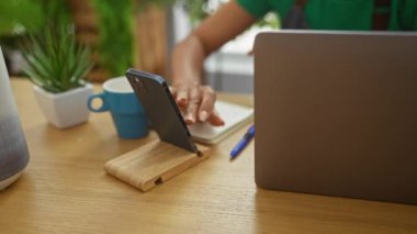 Bitki dolu bir çiçekçide akıllı telefon ve dizüstü bilgisayar kullanan Afrikalı Amerikalı bir kadın.