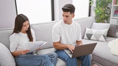 Odaklanmış bir adam ve gülümseyen bir kadın oturma odasındaki koltukta oturmuş kağıtlarla ve bilgisayarla çalışıyor..